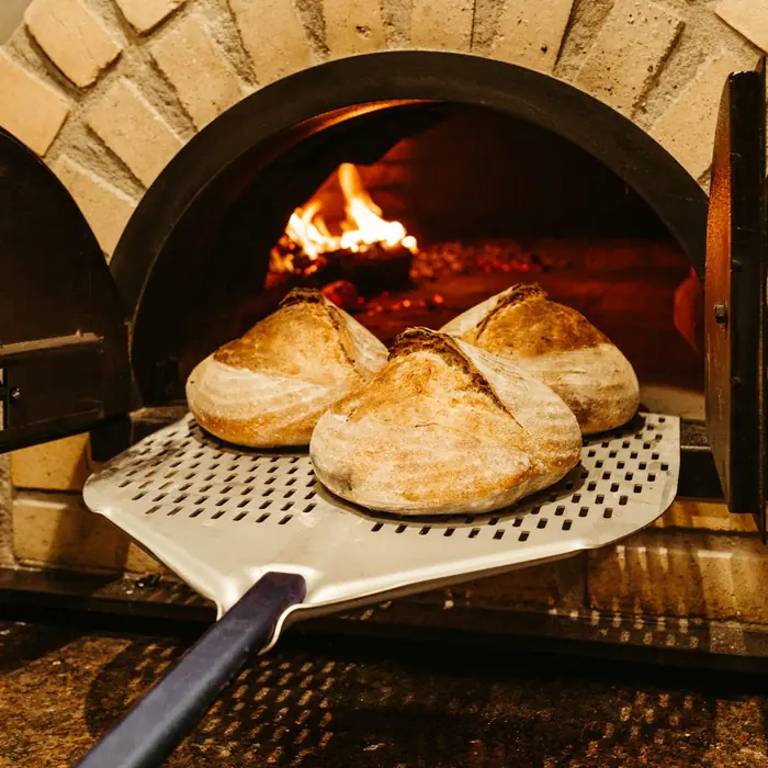 Three scored sourdough loaves on a peel in front of a wood-fired brick oven